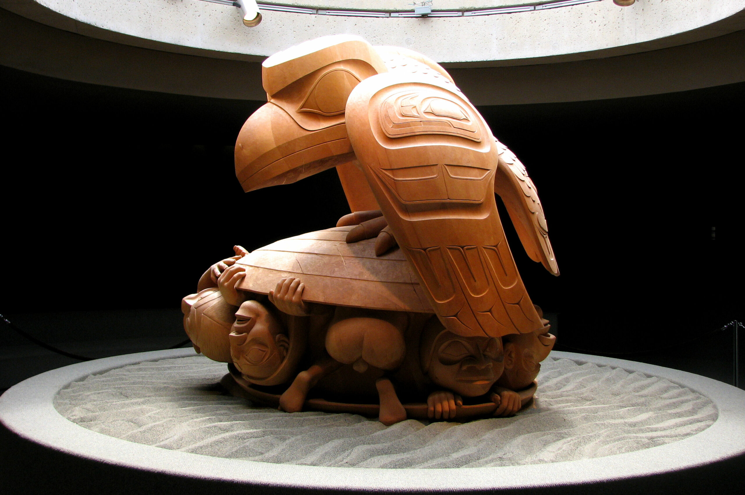 A piece of native artwork: a wood-carved figure of a raven perched atop a clam from which humans emerge.